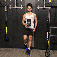 A man leans on a gym wall, holding a Cookies & Cream when protein canister in one hand. The man is wearing a white tank top between two exercise bands.